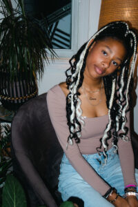 Girl with mixed black and platinum box braids poses in front of some plants.