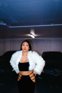 Girl poses in black crop top and black peek-a-boo pants with a fur jacket while standing infront of two mercedes.