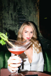 young blonde woman poses wuth cocktail with the glass shaped as a bird while wearing white slong sleeve dress with cleavage.